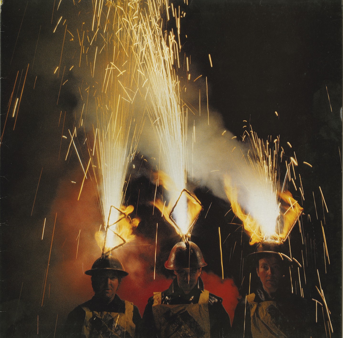 A colour photograph featuring the three members of Bow Gamelan. They are facing the camera and are wearing hemets with sparklers attatched which are shaped to spell out BOW. The sparkers are lit and are firing bright yellow, red and white sparks into the sky above them.